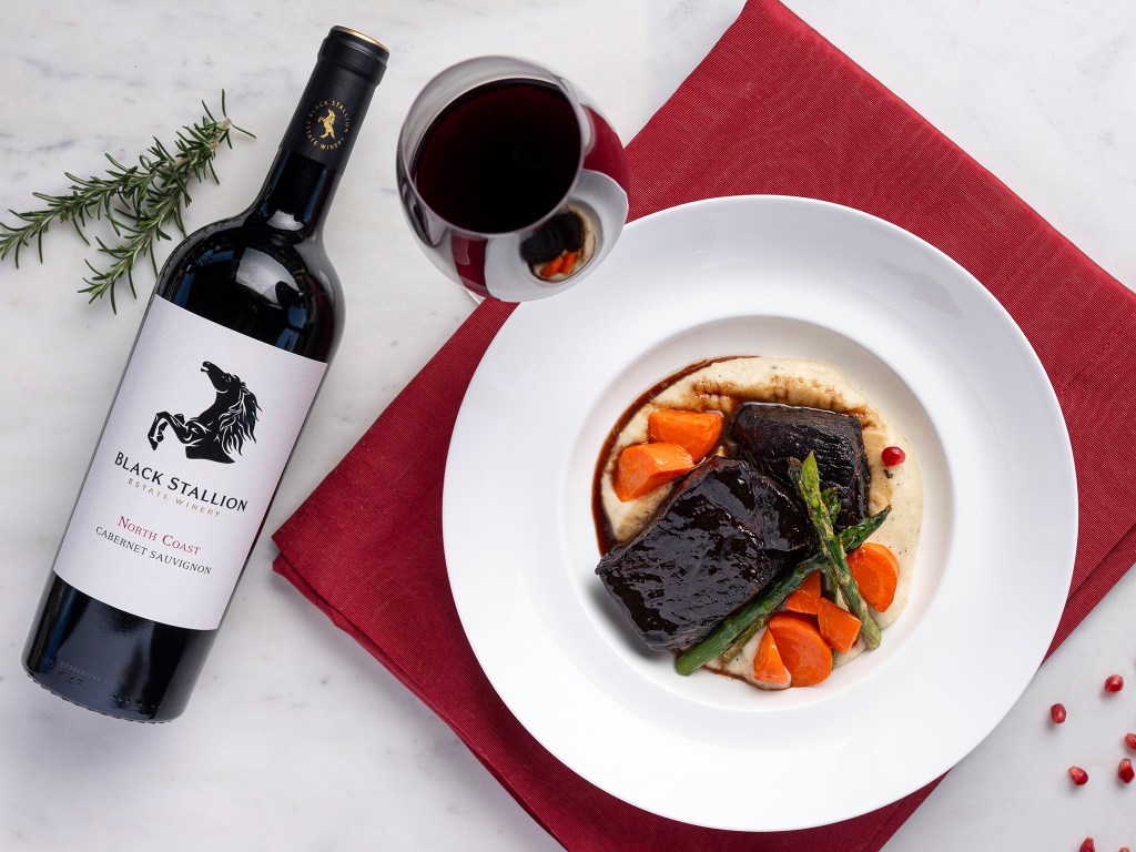 A bottle of Black Stallion Estate Winery North Coast Cabernet Sauvignon next to a glass of wine and a plate of Pomegranate & Cherry Braised Pork